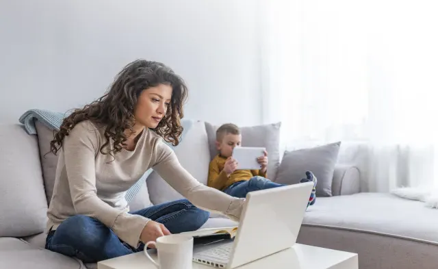 Mother seated on couch taking an online class with son reading on a tablet next to her